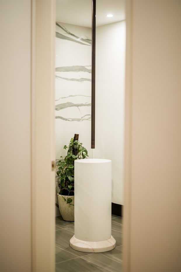 A narrow view through a doorway into a refined bathroom featuring a freestanding cylindrical white basin on a round stone pedestal, a dark suspended tap, large white-and-grey veined wall slabs, dark grey tiled flooring, and a potted plant. The space is softly lit and minimalist with warm neutral tones.