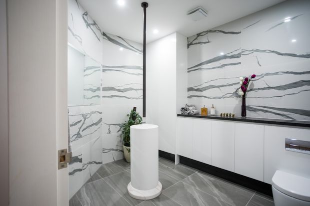 Contemporary bathroom with dramatic white-and-grey veined marble-look wall panels, matching grey floor tiles, a freestanding white basin, wall-mounted white vanity cabinetry, black benchtop and trim, and a toilet. Recessed ceiling lights and a slim black pendant add contrast to the polished, high-end scheme.