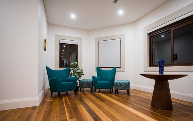 A compact, refined sitting area with two teal upholstered armchairs and matching patterned ottomans arranged around a small side table. The room has warm timber flooring, white walls and ceiling, recessed lighting, roller blinds, and dark timber-framed windows. A sculptural dark wood pedestal table with a blue glass vase sits beside the window.