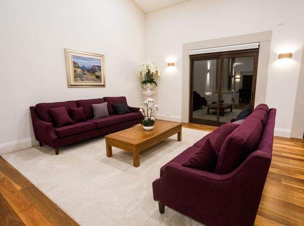 A warm, formal living room with burgundy upholstered sofas arranged around a rectangular timber coffee table. The room has cream walls, a large pale area rug over timber flooring, framed landscape artwork, floral arrangements, wall sconces, and dark-framed glazed double doors with a reflected adjoining room beyond.