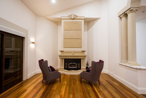 Elegant formal sitting room with a classical fireplace surround and overmantel, paired burgundy upholstered armchairs, ornate columns, warm polished timber flooring, recessed ceiling lighting, and a dark-framed glazed door. The room has a refined, symmetrical composition and traditional detailing.