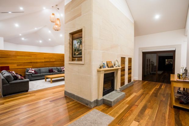Open-plan residential interior showing a living room and adjoining hallway arranged around a large central stone-clad fireplace. The living area has a dark sectional sofa, timber feature wall, coffee table, and decorative pendant lights, while the surrounding space features polished warm timber flooring, white walls and ceiling, recessed lighting, and dark timber-framed openings.