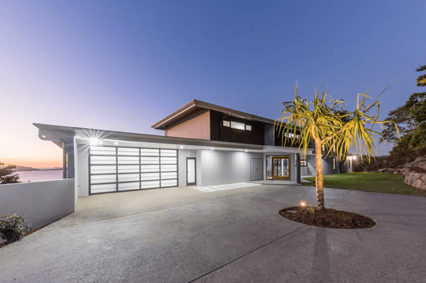 Exterior twilight view of a contemporary two-storey residence on a sloping site. The home features a broad attached garage with translucent horizontal-panel doors, a flat-roofed lower wing, dark upper-level cladding, extensive glazing, a concrete driveway, landscaped lawn, and a palm-like tree in a circular planting bed. Water and distant landscape are faintly visible beyond the property.