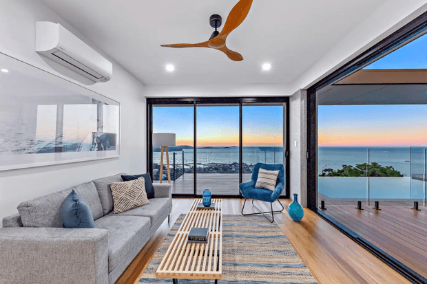 Bright contemporary living room with a large grey sectional sofa, patterned cushions, blue accent chair, slatted timber coffee table, and woven rug. Full-height glazing opens onto a timber balcony with glass balustrades and expansive ocean views. A timber-bladed ceiling fan, recessed ceiling lights, air-conditioning unit, and minimal decor complete the relaxed coastal setting.