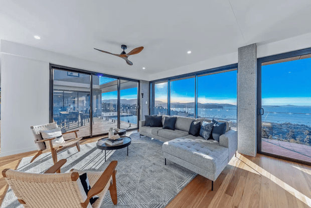 Bright contemporary living room with expansive floor-to-ceiling glazing overlooking a coastal waterfront and cityscape. The room features a large light-grey sectional sofa, woven timber lounge chairs, round nesting coffee tables, a geometric grey area rug, timber flooring, exposed concrete columns, and a timber-bladed ceiling fan. Strong natural light and panoramic views create an airy, refined atmosphere.