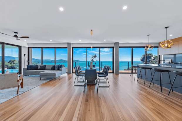 Open-plan contemporary coastal living space with floor-to-ceiling glazing overlooking a blue waterfront and distant hills. The room combines a living area, dining table, and sleek kitchen with island seating. Warm timber flooring, dark grey cabinetry and furnishings, exposed concrete columns, and geometric pendant lights create a refined modern interior.