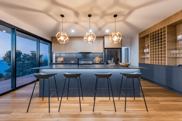 A spacious contemporary kitchen with a long dark island and four bar stools, extensive floor-to-ceiling glazing along one side, dark integrated cabinetry, a timber wine-grid display, and three geometric pendant lights. Warm timber flooring contrasts with the charcoal joinery and cool blue-grey benchtop surfaces. A landscape is visible through the glazing.