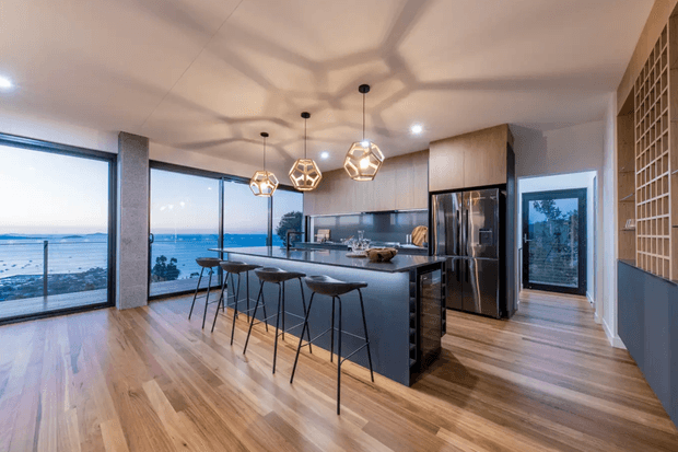 Contemporary open-plan kitchen with a large dark island and bar seating, stainless-steel appliances, extensive floor-to-ceiling glazing, and a warm timber floor. Geometric pendant lights and a timber grid-screen feature add visual interest. The glazing opens to a broad coastal or waterfront view.