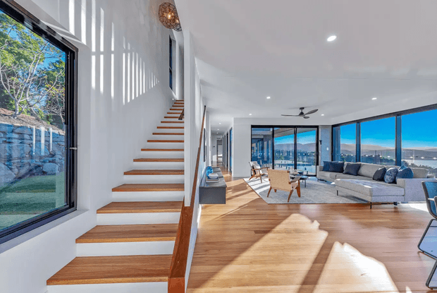 Bright, open-plan contemporary living area with a timber staircase, expansive floor-to-ceiling glazing, and panoramic water and landscape views. The room features warm timber flooring, white walls and ceiling, a large neutral sectional sofa, and minimalist furnishings. Strong natural light creates a spacious, airy atmosphere.