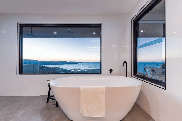 Modern minimalist bathroom featuring a freestanding white bathtub positioned beside large black-framed corner windows overlooking a coastal or waterfront landscape. The room has pale glossy wall surfaces, large grey floor tiles, a simple dark floor-mounted bath filler, and a calm, airy atmosphere.