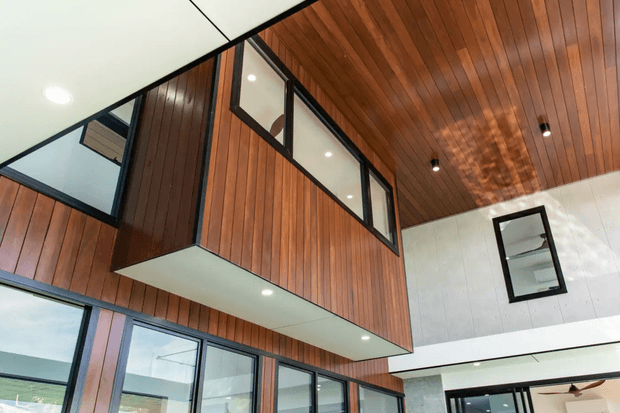 Interior architectural view of a contemporary double-height space featuring extensive warm vertical timber wall and ceiling lining, black-framed clerestory glazing, a projecting upper-level timber-clad volume, recessed downlights, and contrasting white wall panels. The lower level includes large glazed doors or windows, creating an open, airy connection between levels.