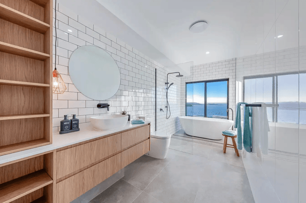 Bright contemporary bathroom with a freestanding white bathtub positioned beside expansive windows overlooking water. The room features white subway-tile walls, large grey floor tiles, a floating timber vanity with vessel basin, round frameless mirror, open timber shelving, wall-mounted black tapware and shower fittings, and a glass shower screen. A stool and folded towels add light styling.