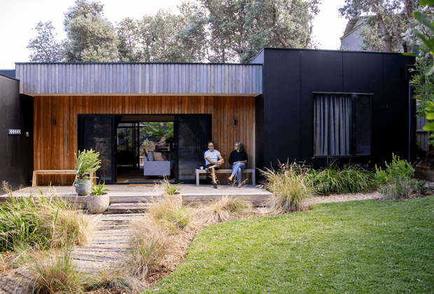 Front exterior view of a contemporary single-storey home set within a landscaped garden. The facade combines dark painted cladding with warm vertical timber lining around a recessed entry and glazed living area, plus a corrugated metal roof and large dark-framed openings. A stepping-stone path crosses ornamental grasses and lawn toward the covered porch, where two people are seated.
