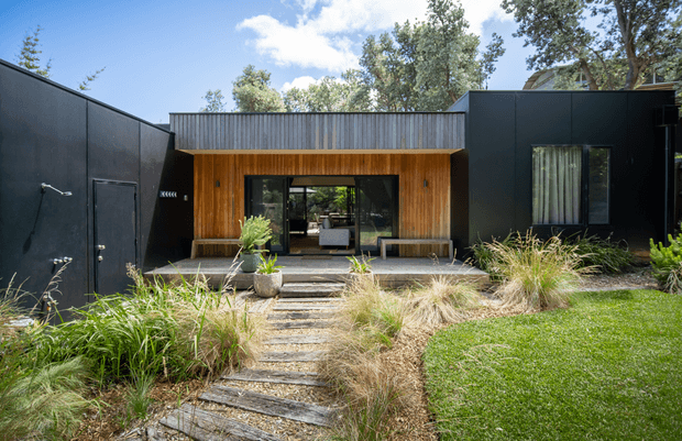 Front exterior view of a contemporary single-storey home with bold matte black cladding, a central recessed entry clad in warm timber, extensive glazing, and a corrugated metal roof or parapet. A stepping-stone path leads through a landscaped garden of ornamental grasses to the entrance.