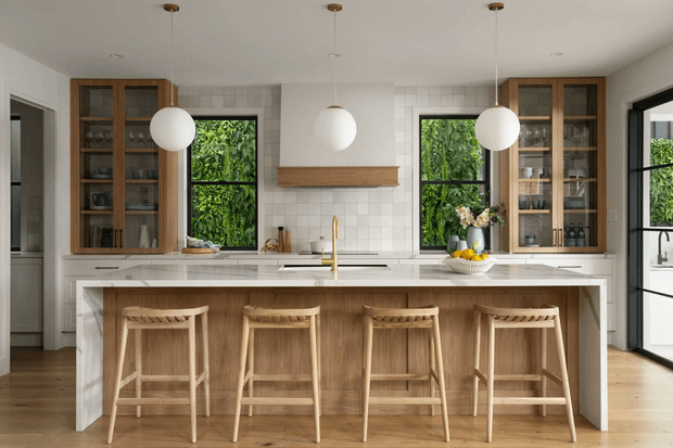 Bright, spacious contemporary kitchen with a large white stone island and four timber bar stools. The kitchen features warm timber cabinetry with glass-front display doors, a white tiled splashback, integrated appliances, a timber rangehood shelf, brass-toned tapware, black-framed windows and doors, and three spherical pendant lights. Lush greenery is visible through the windows.
