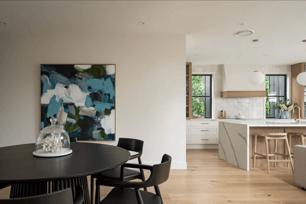 Open-plan contemporary dining and kitchen interior. A black round dining table with dark chairs occupies the foreground, separated from a bright kitchen by a partial wall. The kitchen features light timber flooring, white cabinetry, a pale stone island with subtle veining, a tiled splashback, black-framed windows, timber shelving, and pendant lighting. An abstract artwork and glass centrepiece add visual interest.