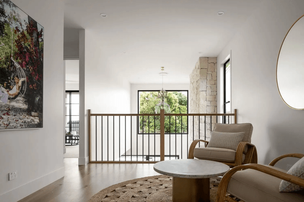 Bright, refined contemporary interior lounge or landing with pale timber flooring, white walls and ceiling, a natural stone feature wall, timber-and-black-metal balustrade, rattan lounge chairs, a round stone-topped coffee table, and a large window overlooking greenery. A stair or void railing and adjacent open spaces suggest an upper-level living or circulation area.
