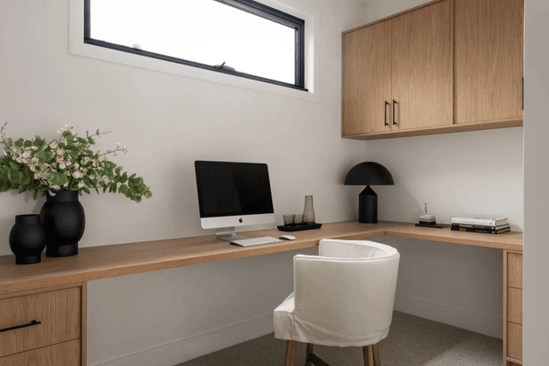 A compact, calm home office with a wall-to-wall timber desk, integrated drawers and overhead cabinetry. A computer, black dome desk lamp, books and dark vases sit on the desk, with a white upholstered chair in the foreground. The room has warm wood joinery, neutral painted walls, a high horizontal window and carpeted flooring.