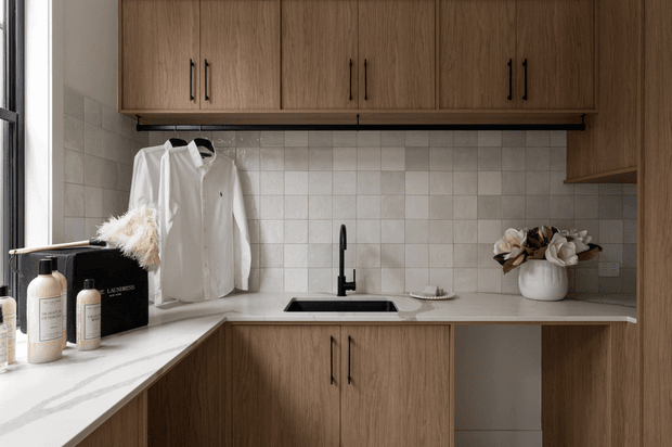 Compact contemporary laundry with warm wood-grain overhead and base cabinetry, a white stone-look benchtop, square light tiled splashback, and a black undermount sink with matching tap. A hanging white shirt, laundry products, and a small vase add domestic context. Natural light enters from the left.