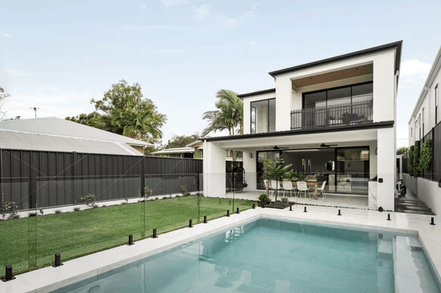 A contemporary two-storey residence viewed from the backyard, with a large rectangular swimming pool in the foreground, manicured lawn, dark fencing, and a covered outdoor entertaining area beneath the upper level. The home has a crisp white rendered facade, dark metal-framed glazing and balustrades, a shallow sloping roofline, and broad indoor-outdoor openings connecting to a dining and living space. Tropical planting is visible around the suburban property.
