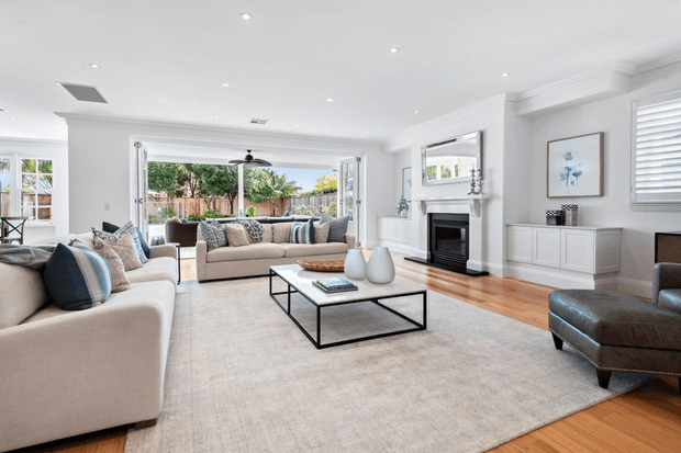 A bright, spacious living room with white walls, recessed lighting, built-in cabinetry, and a traditional fireplace with a white mantel. Two large neutral sofas with blue and patterned cushions face a black metal-framed coffee table over a large light rug. Timber flooring, plantation shutters, framed artwork, and wide folding glass doors opening to a landscaped outdoor area complete the refined interior.