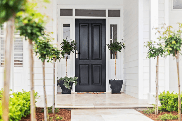 Bright residential front entry with a black paneled door framed by white trim and sidelights, set within a white horizontal weatherboard facade. Symmetrical potted topiary trees and low hedging line the pale stone or concrete porch and walkway.