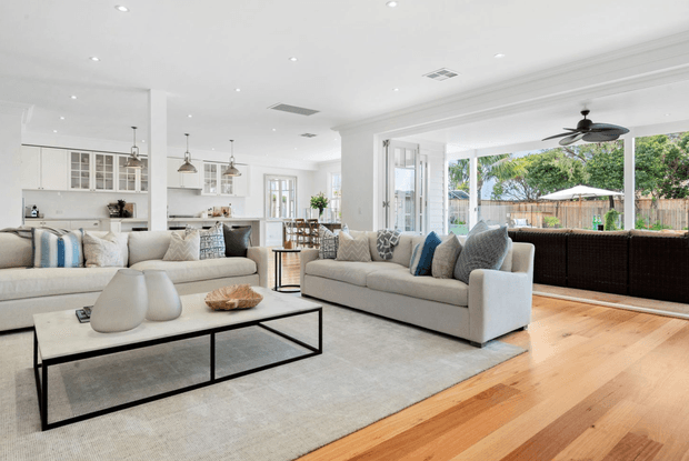 Bright, open-plan contemporary living area with two large upholstered sofas, a low black-framed coffee table, neutral rug, and blue-grey cushions. The space flows into a white kitchen and dining area, with extensive glazing opening toward a leafy outdoor setting. Light timber flooring, white walls, recessed lighting, and a ceiling fan create an airy, refined family-home interior.