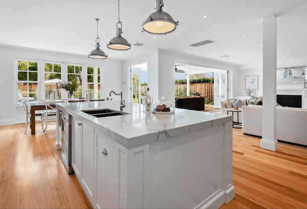 Bright open-plan kitchen centred on a large white island with a stone benchtop, undermount sink and polished tapware. White shaker-style cabinetry, warm timber flooring, white walls and several industrial-inspired pendant lights define the kitchen, which opens to a dining area and living room with an outdoor space visible beyond.