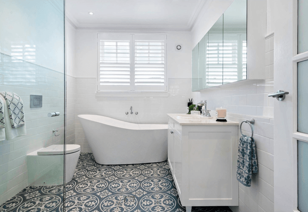 Bright, refined bathroom featuring a freestanding white bathtub beneath plantation shutters, a glass-enclosed shower, wall-mounted vanity with integrated basin, mirrored cabinetry, white subway-tile walls, and patterned encaustic-style floor tiles. The palette is predominantly white and grey-blue with decorative dark patterned flooring.