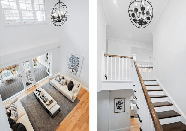 Split-view interior photograph showing a bright, high-ceilinged living room and adjacent stairway. The living area has white walls, large multi-pane windows, pale timber flooring, two light sofas, a large grey rug, and a dark linear coffee table. The second view shows a white stairwell with dark timber stair treads, a polished timber handrail, white balustrades, decorative wall mouldings, and black spherical cage chandeliers.