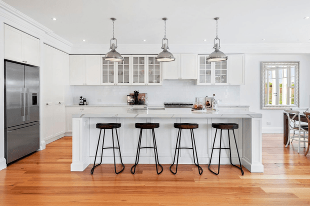 Bright, spacious white kitchen with a large central island and four counter stools. The room features white shaker-style cabinetry, glass-front upper cabinets, a pale stone benchtop and splashback, stainless-steel refrigerator, polished timber flooring, and three industrial-style pendant lights. A dining area is visible through the opening at right.
