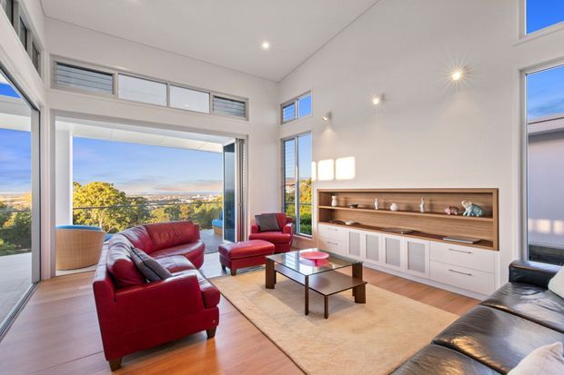 Bright, spacious contemporary living room with high ceilings, extensive floor-to-ceiling glazing, and wide views over the surrounding landscape. The room features warm timber flooring, white walls, a built-in timber and white storage unit, red leather lounge chairs and sofa, a glass-top coffee table, and a large neutral rug.