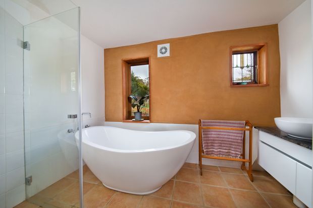 Bright contemporary bathroom featuring a freestanding white bathtub, frameless glass shower enclosure, floating white vanity with round vessel basin, terracotta-toned plaster feature wall, timber-framed windows, and terracotta floor tiles. A wooden towel stand adds warmth to the otherwise clean, minimal space.
