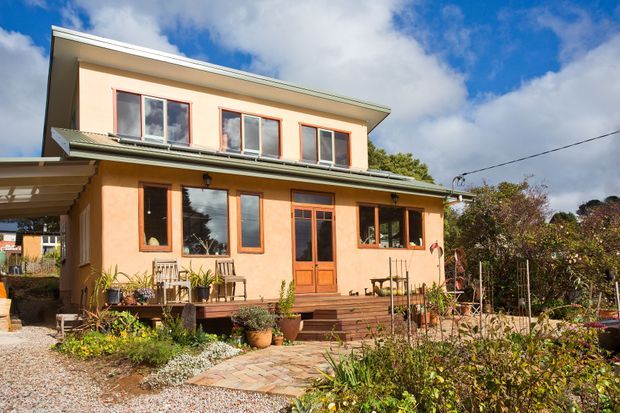 Two-storey residential house with warm peach rendered walls, contrasting timber-framed windows and doors, a shallow low-pitched roof, and a timber verandah/deck. The home is viewed externally from a landscaped garden with a paved path, planting, and a covered side porch. Bright daylight and blue sky create a welcoming suburban residential setting.