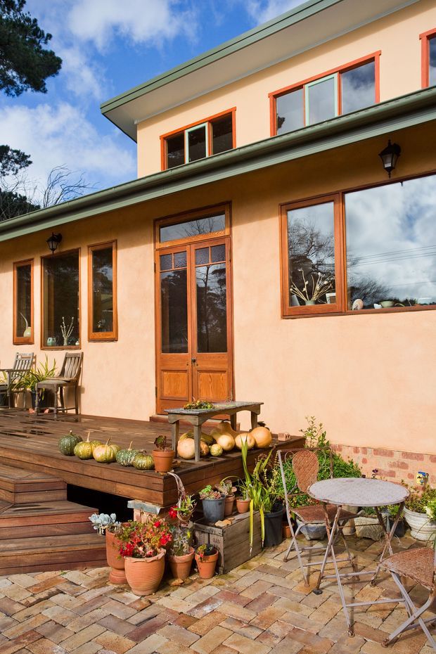 A warm, sunlit exterior view of a two-storey rendered house with green metal roof edging, orange timber-framed windows and doors, and a covered veranda. A timber deck with steps leads to a paved patio furnished with a small round table and chairs, surrounded by potted plants, garden produce, and built-in raised planters.