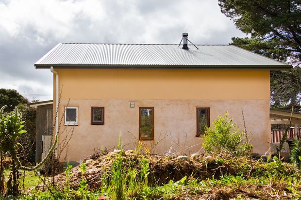 A small single-storey rendered house viewed from the garden, with a simple rectangular facade, shallow corrugated metal gable roof, narrow timber-framed windows, and a warm ochre upper wall over a weathered pale-rendered lower wall. Established garden planting and a covered side structure are visible around the building.