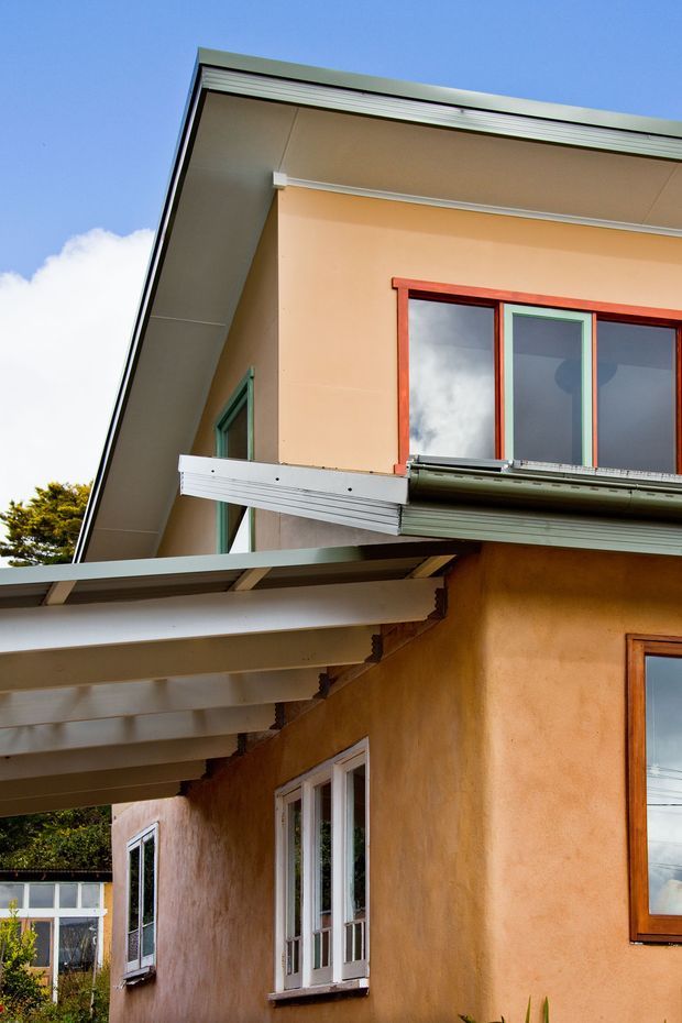 Close-up exterior view of a two-storey house with warm ochre rendered walls, projecting roof and awnings, and contrasting timber-framed windows. The image emphasizes the junction of the rooflines, eaves, metal flashing, and facade detailing under a clear blue sky.