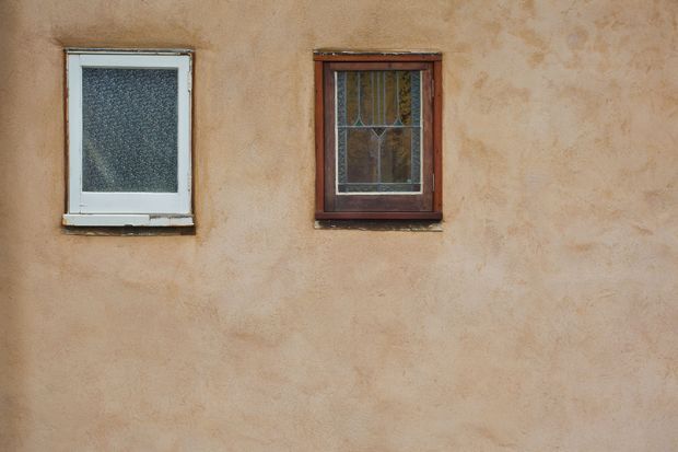Close-up of a textured warm beige stucco exterior wall with two contrasting windows: a white-framed frosted-glass window and a dark timber-framed decorative leaded-glass window.