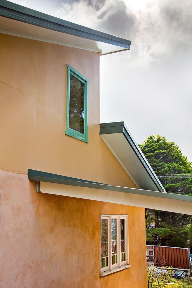 Exterior view of a two-storey rendered building with warm ochre walls, dark green roof edging and projecting eaves, and contrasting turquoise-green window frames. The image highlights the rooflines, facade junctions, doors, and architectural detailing, with mature trees and a small deck or garden area visible around the building.