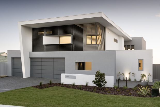 Contemporary two-storey residential facade with crisp white and grey rendered walls, a flat roof, integrated garage, upper-level horizontal louvres, and a minimalist landscaped front garden. The street-facing elevation combines strong geometric volumes with sheltered glazing and warm interior light visible through windows.