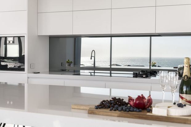 Bright contemporary white kitchen with extensive handleless cabinetry, a large island, integrated appliances, dark stone or glass splashback, and a wide ocean-facing window. A black gooseneck tap, cooktop, and sink are positioned along the rear bench, while the island is styled with a timber serving board, fruit, wine, and glassware.