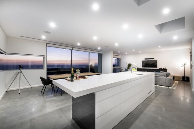 Open-plan contemporary interior dominated by a large white kitchen island, with white handleless cabinetry, polished concrete flooring, expansive windows, and an adjoining living area. A dining table and chairs sit near the glazing, while the living room includes a grey sofa and wall-mounted television. The space has recessed downlights and views toward the ocean or distant landscape at dusk.