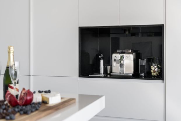 A clean, contemporary kitchen coffee station built into a wall of handleless white cabinetry. The recessed niche has a glossy black interior and contains a coffee machine, grinder, milk frother and glassware. In the foreground, a pale stone island or benchtop holds a timber serving board with champagne, fruit and cheese.