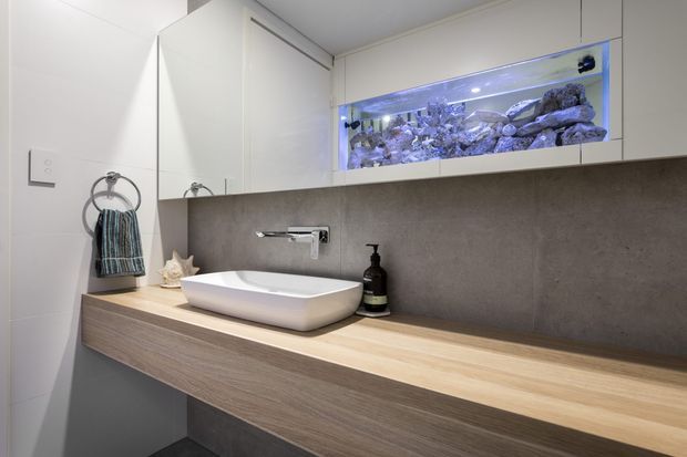Contemporary bathroom vanity with a white vessel basin set on a long light timber benchtop, wall-mounted chrome tapware, white cabinetry, and a dark grey textured splashback. An illuminated built-in aquarium forms a distinctive feature above the vanity. The space is compact, clean, and softly lit.