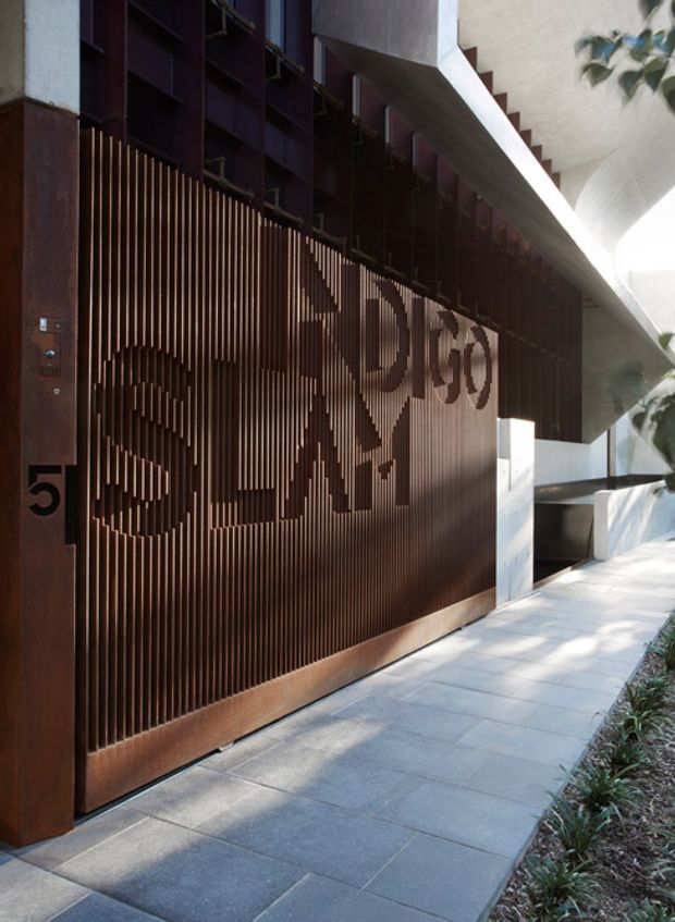 Exterior architectural view of a contemporary multi-storey building entrance. A tall rust-toned metal screen and gate with vertical slats incorporates large cut-out lettering and the number 5. Dark structural elements, a projecting white concrete canopy, and a paved pedestrian walkway with landscaped planting create a dramatic, sculptural facade.