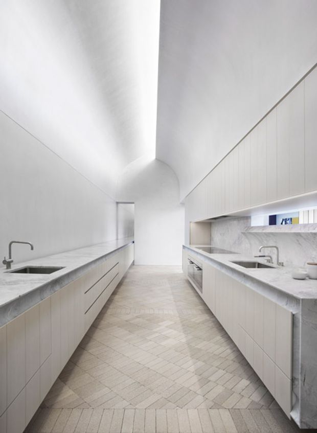 A bright, minimalist galley kitchen with two long parallel runs of pale cabinetry and substantial light stone benchtops. Integrated sinks and brushed metal gooseneck taps sit on both sides, while a marble-look stone splashback and concealed under-cabinet lighting define the right-hand joinery. The room has a high, sculptural curved ceiling transition at the far end, white walls, and a pale herringbone/parquet-style tiled floor.
