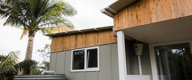 Exterior view of a contemporary low-rise building with vertical timber cladding, muted grey-green wall panels, dark-framed windows, and a covered entry or porch supported by a white column. Palm trees and lush vegetation surround the structure, suggesting a warm landscaped setting.