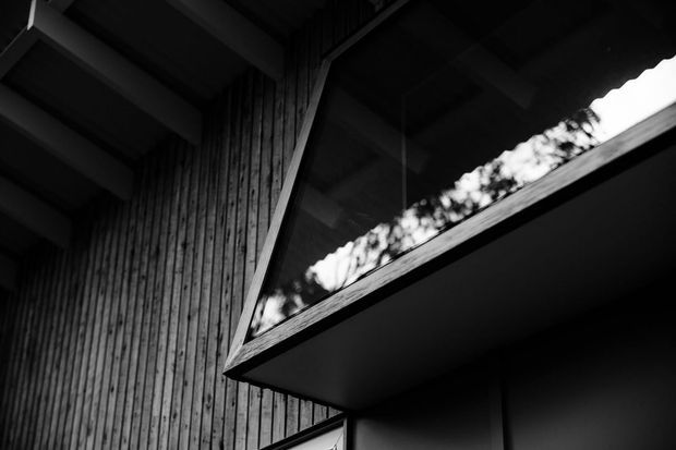 Black-and-white architectural detail showing a sharply angled roof or canopy junction, dark metal or timber framing, vertical timber wall cladding, and reflective glazing beneath an overhanging roofline. The high-contrast composition emphasizes geometric forms and construction details.