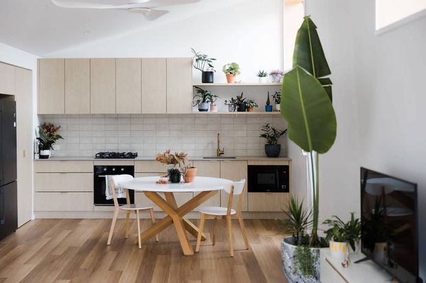 Bright open-plan kitchen and dining area with light timber cabinetry, white tiled splashback, timber flooring, a compact dining table and chairs, and abundant indoor plants. A living area with television is visible at right. The space has a clean, contemporary Scandinavian-inspired appearance with soft neutral finishes and natural materials.