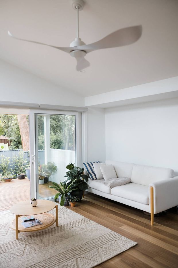 Bright, compact living room with white walls and ceiling, a sculptural white ceiling fan, pale upholstered sofa, indoor plants, layered round wooden coffee tables, and a textured neutral rug. Warm timber flooring continues toward open glazed doors leading to a timber outdoor deck and garden. The room has a clean, airy contemporary aesthetic with abundant natural light.